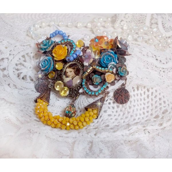 Broche Eclats P&eacute;tillants d&rsquo;inspiration Baroque, orn&eacute;e de fleurs en r&eacute;sine autour d&rsquo;un cabochon strass&eacute;s et perl&eacute;s sur estampes, cristaux, breloques, perle cloisonn&eacute;e, perles jaunes en verre et roses. 