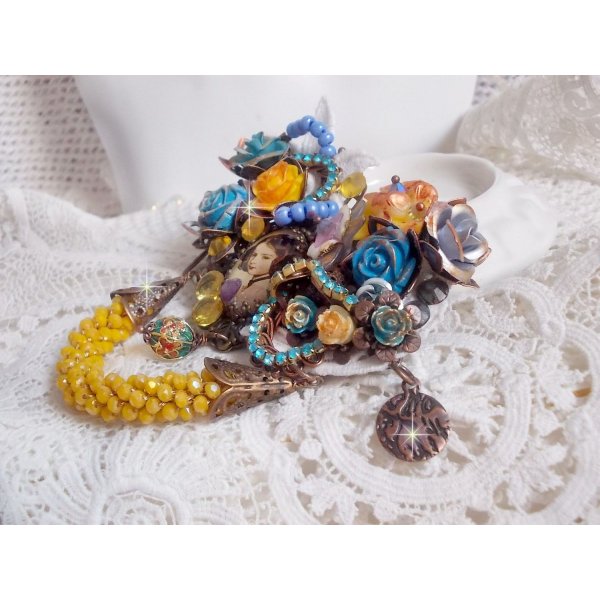 Broche Eclats P&eacute;tillants d&rsquo;inspiration Baroque, orn&eacute;e de fleurs en r&eacute;sine autour d&rsquo;un cabochon strass&eacute;s et perl&eacute;s sur estampes, cristaux, breloques, perle cloisonn&eacute;e, perles jaunes en verre et roses. 