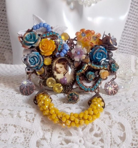 Broche Eclats P&eacute;tillants d&rsquo;inspiration Baroque, orn&eacute;e de fleurs en r&eacute;sine autour d&rsquo;un cabochon strass&eacute;s et perl&eacute;s sur estampes, cristaux, breloques, perle cloisonn&eacute;e, perles jaunes en verre et roses. 